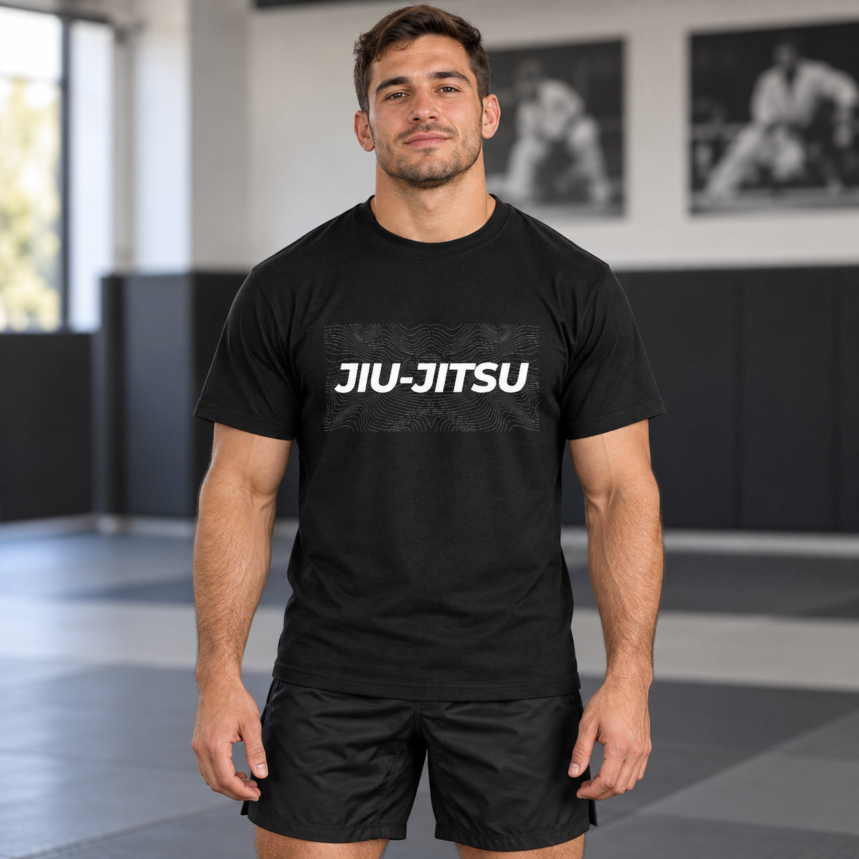 A man wearing a black short-sleeve t-shirt with 'JIU-JITSU' printed in white across the chest, standing in a gym with black mats and framed black-and-white photos on the wall.